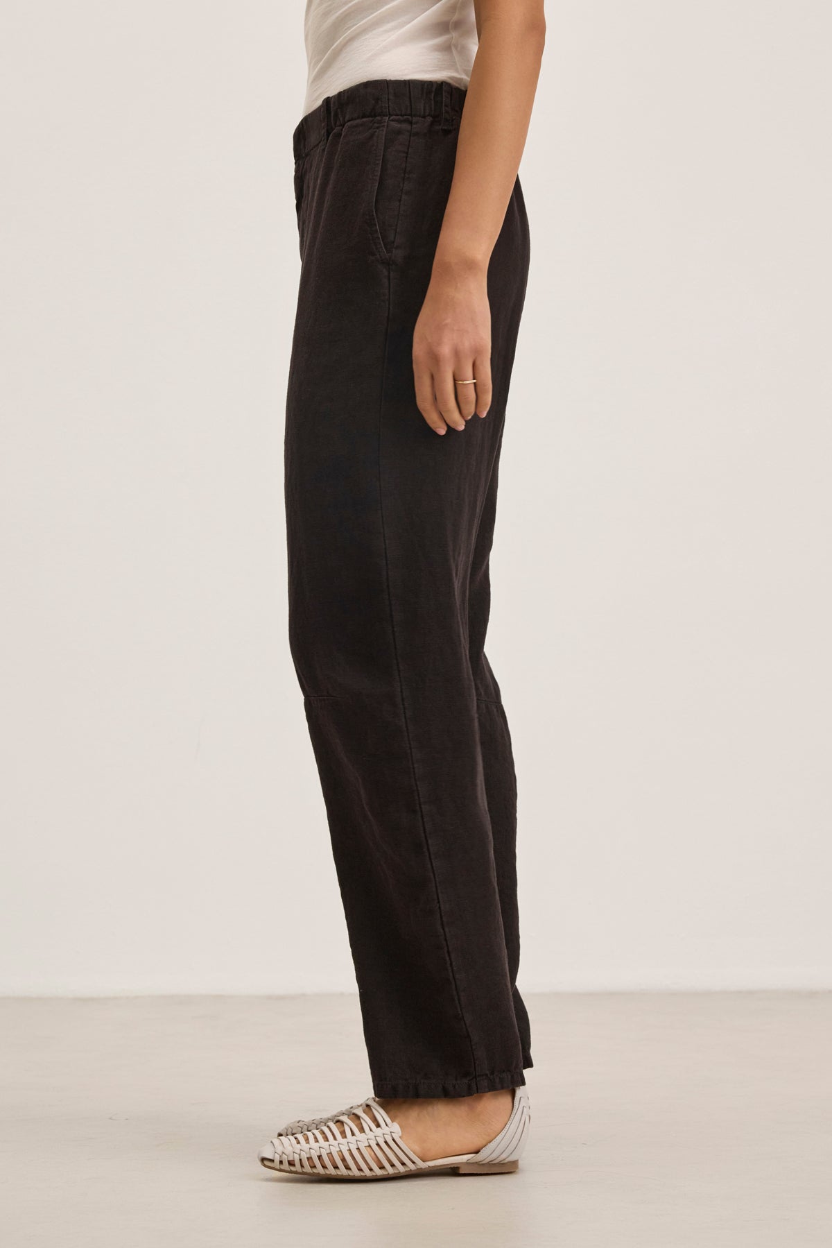 A person stands sideways against a plain light background, wearing a white top, TAYLOR HEAVY LINEN PANT by Velvet by Graham & Spencer in black, and white woven flat shoes.-39111849214145