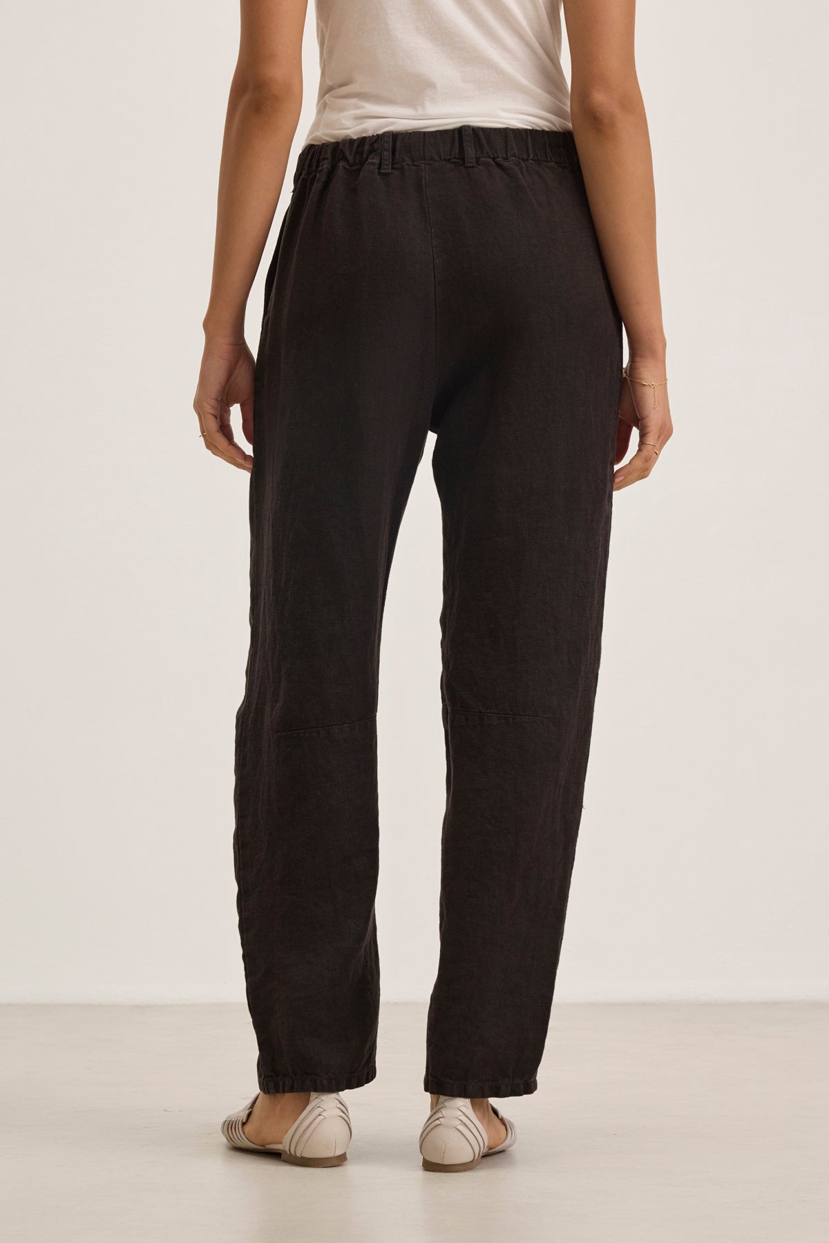 A person stands facing away, wearing a white short-sleeve top and the Velvet by Graham & Spencer TAYLOR HEAVY LINEN PANT in black with an elastic waistband, paired with beige flats, against a plain light background.-39111850000577
