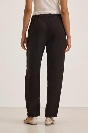 A person stands facing away, wearing a white short-sleeve top and the Velvet by Graham & Spencer TAYLOR HEAVY LINEN PANT in black with an elastic waistband, paired with beige flats, against a plain light background.