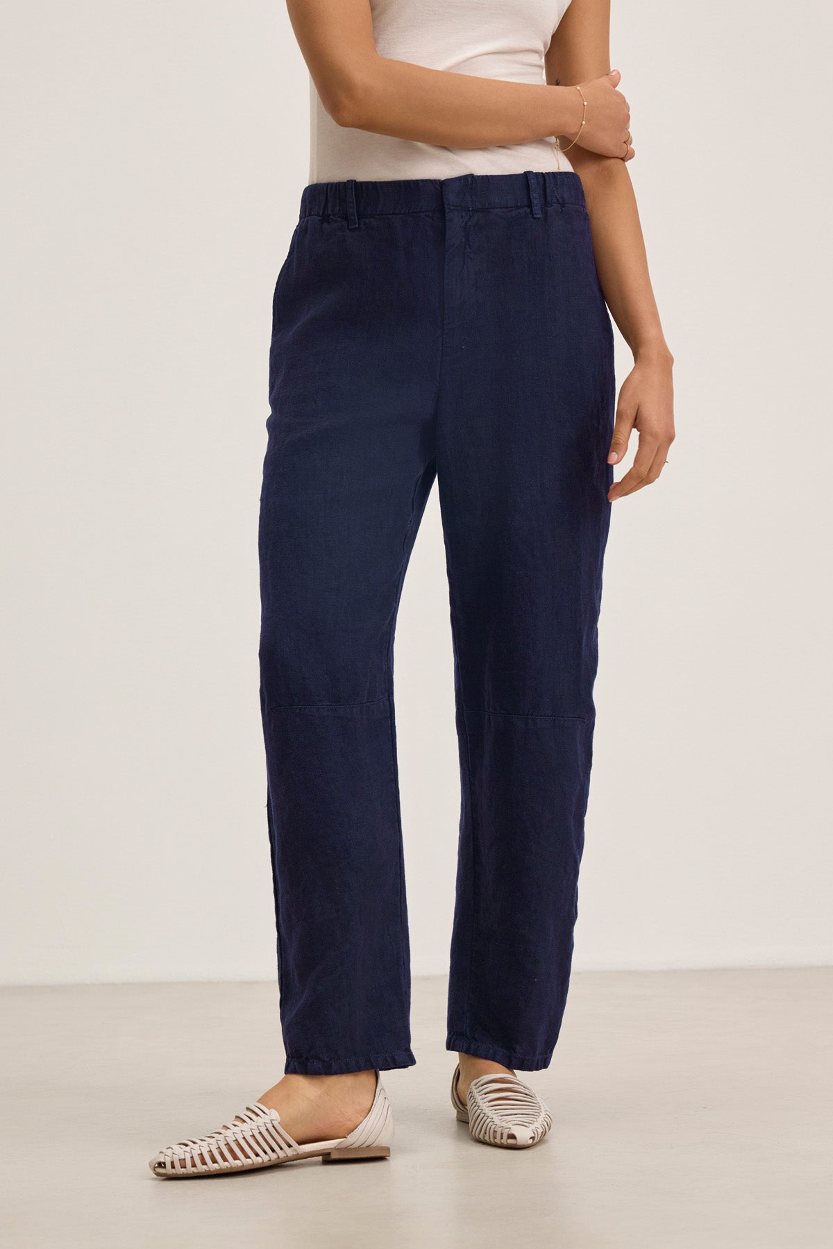 A person stands against a plain background wearing the TAYLOR HEAVY LINEN PANT by Velvet by Graham & Spencer in navy blue, paired with a white sleeveless top and white woven flat shoes.-41645170360513
