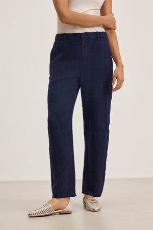 A person stands against a plain background wearing the TAYLOR HEAVY LINEN PANT by Velvet by Graham & Spencer in navy blue, paired with a white sleeveless top and white woven flat shoes.