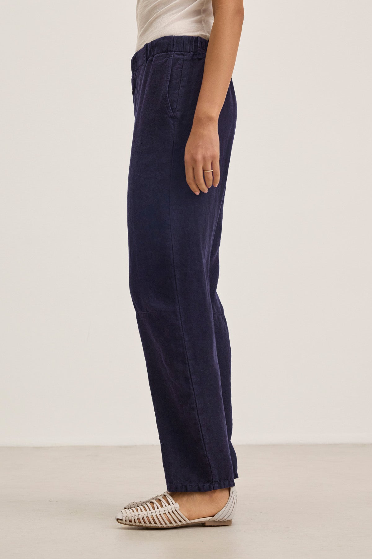 A person stands sideways against a plain light background, wearing Velvet by Graham & Spencer's TAYLOR HEAVY LINEN PANT in navy blue with a barrel leg, paired with a white short-sleeve top and white woven flat sandals.