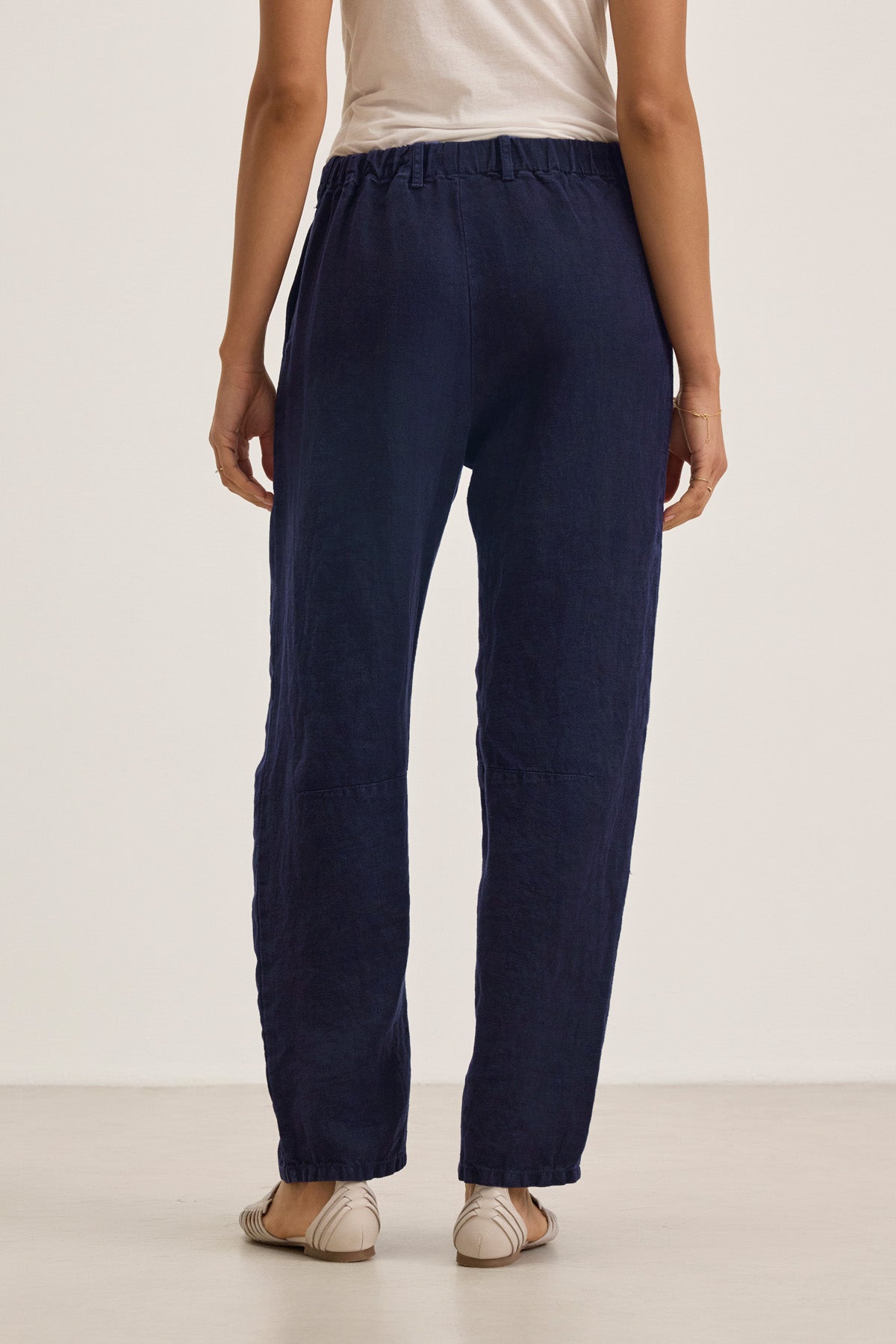 A person stands facing away, wearing a white top, beige flat shoes, and Velvet by Graham & Spencer's TAYLOR HEAVY LINEN PANT in navy blue with barrel legs and an elastic waistband.-41645170426049