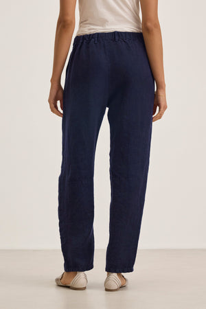 A person stands facing away, wearing a white top, beige flat shoes, and Velvet by Graham & Spencer's TAYLOR HEAVY LINEN PANT in navy blue with barrel legs and an elastic waistband.