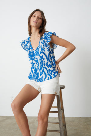 Woman in a ALEAH PRINTED COTTON GAUZE TOP by Velvet by Graham & Spencer and white shorts posing with hand on hip, leaning on a wooden stool.