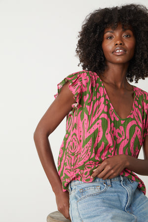 A black woman is sitting on a stool wearing a Velvet by Graham & Spencer ALEAH PRINTED COTTON GAUZE TOP in pink and green.