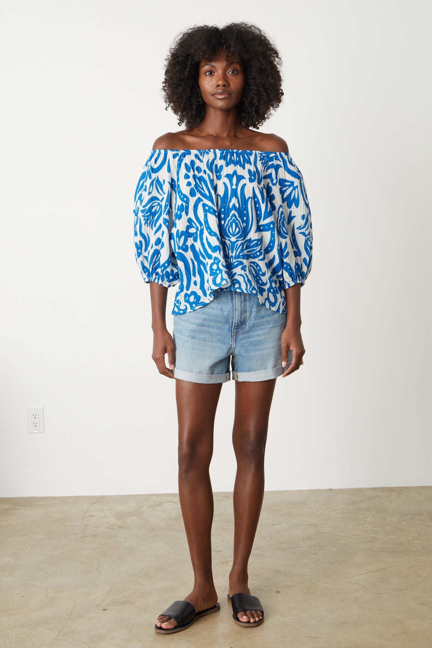 A woman wearing a Velvet by Graham & Spencer CANDICE PRINTED COTTON GAUZE TOP in bold blue and white print, off shoulder, with denim shorts full length front with sandals-26577386832065