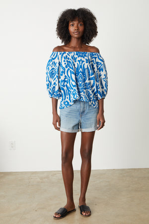 A woman wearing a Velvet by Graham & Spencer CANDICE PRINTED COTTON GAUZE TOP in bold blue and white print, off shoulder, with denim shorts full length front with sandals