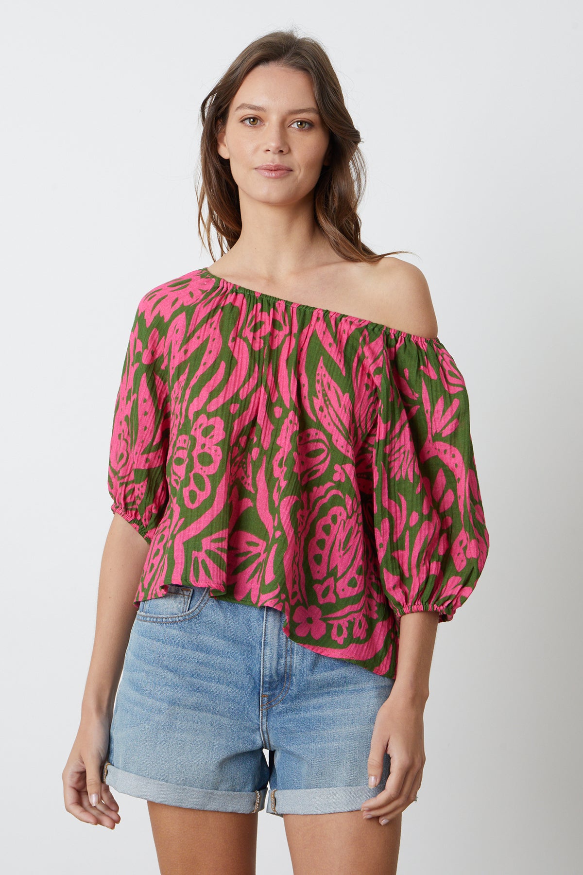   A model wearing a Velvet by Graham & Spencer CANDICE PRINTED COTTON GAUZE TOP in bold pink and green print off shoulder with denim shorts front 