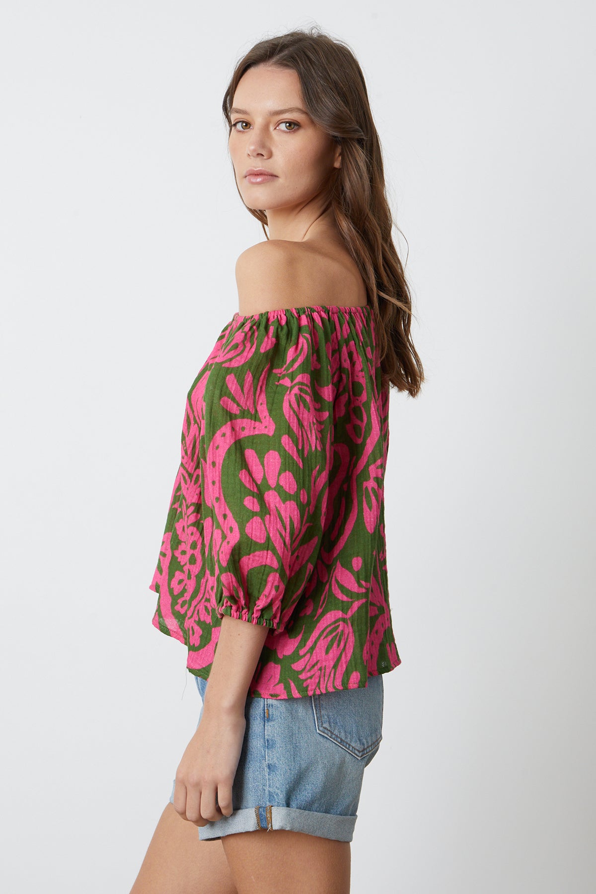   The model is wearing a pink and green off the shoulder CANDICE PRINTED COTTON GAUZE TOP by Velvet by Graham & Spencer with denim shorts, side view. 