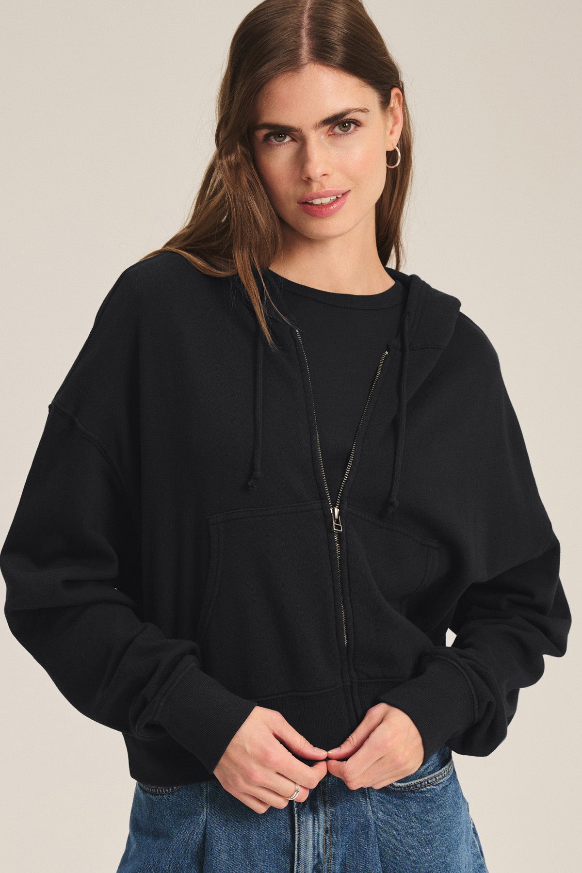A woman with long brown hair models the HIGHLAND HOODIE by Velvet by Graham & Spencer—a black fleece hoodie with a drawstring hood—paired with blue jeans, standing against a plain background.-40293783240897