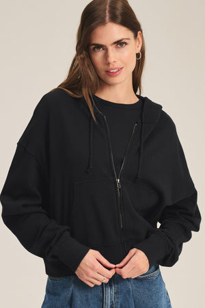 A woman with long brown hair models the HIGHLAND HOODIE by Velvet by Graham & Spencer—a black fleece hoodie with a drawstring hood—paired with blue jeans, standing against a plain background.