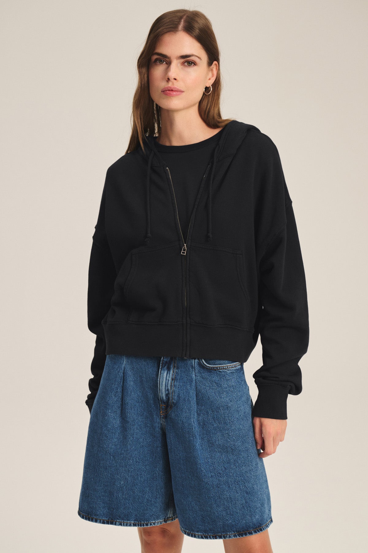 A woman models the HIGHLAND HOODIE by Velvet by Graham & Spencer—a black fleece hoodie with a drawstring hood—paired with blue denim shorts, set against a plain, light background.