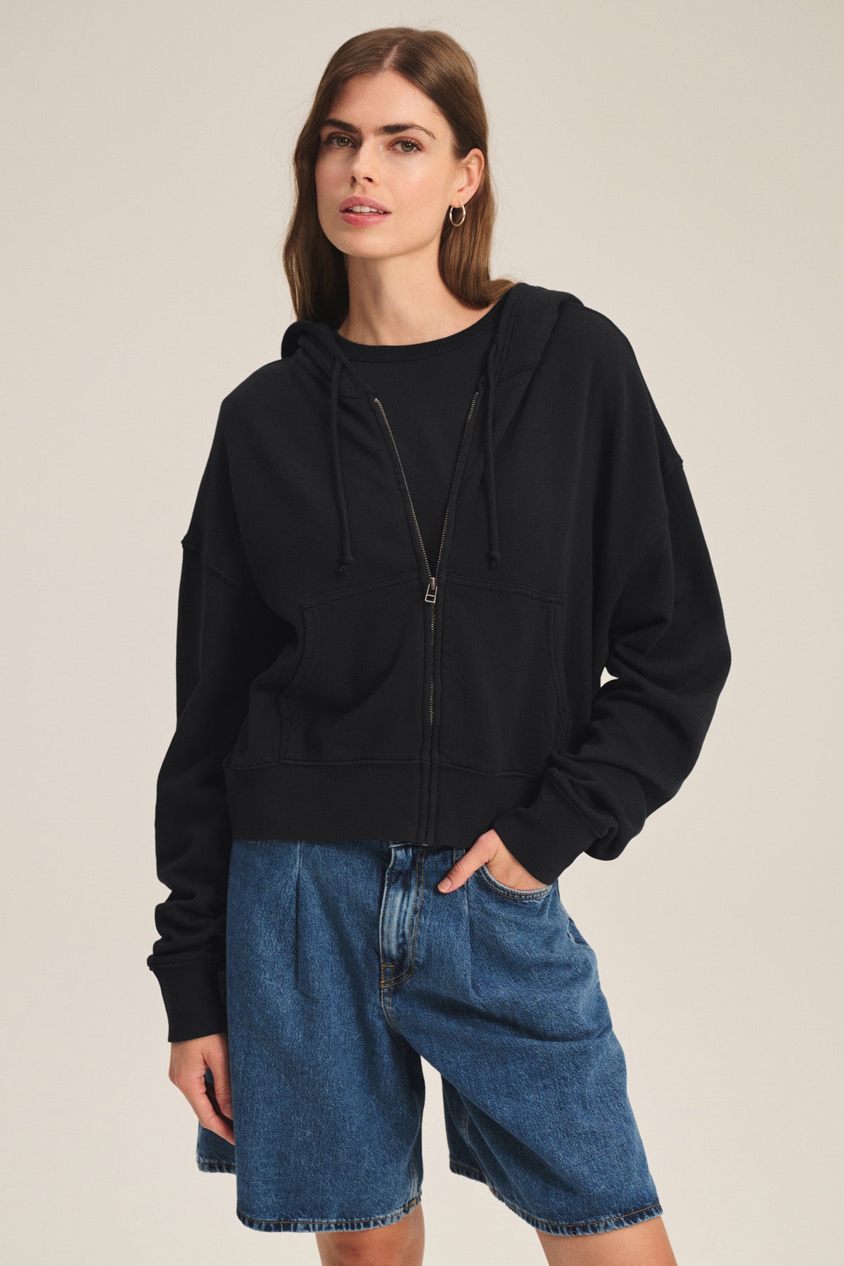 A woman models the HIGHLAND HOODIE by Velvet by Graham & Spencer, a black fleece hoodie with a drawstring hood, layered over a black shirt and paired with blue denim shorts, standing with one hand in her pocket against a plain background.