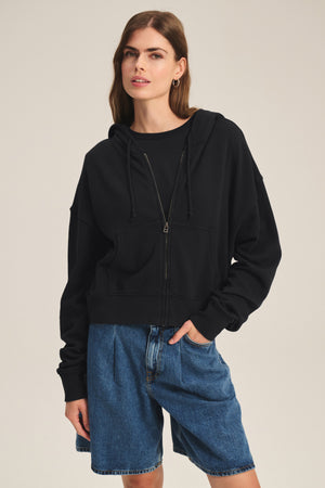 A woman models the HIGHLAND HOODIE by Velvet by Graham & Spencer, a black fleece hoodie with a drawstring hood, layered over a black shirt and paired with blue denim shorts, standing with one hand in her pocket against a plain background.