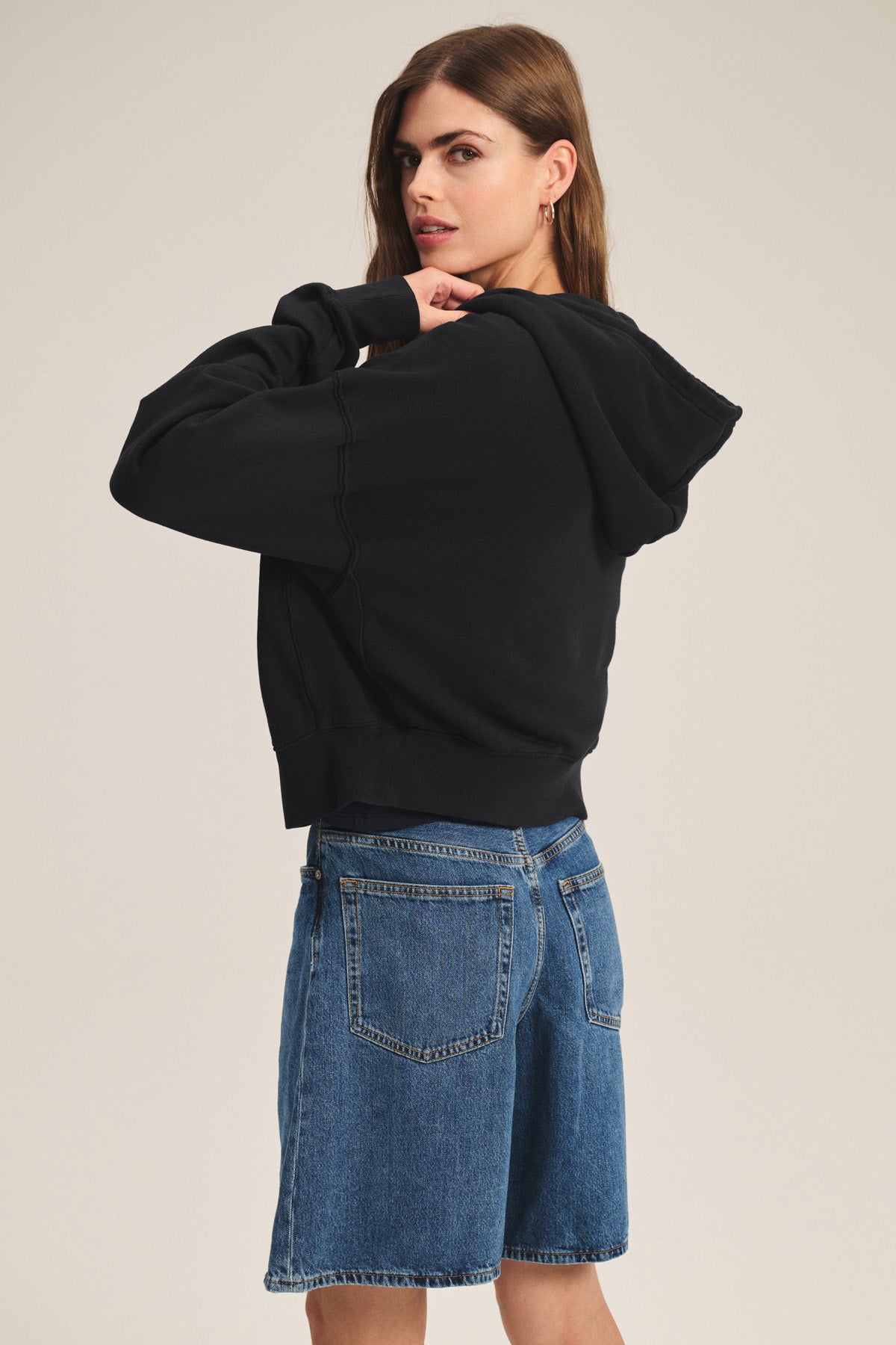 Wearing the HIGHLAND HOODIE by Velvet by Graham & Spencer, this person stands sideways in a black full zip hoodie with drawstring hood and blue denim shorts, glancing over their shoulder against a plain background.