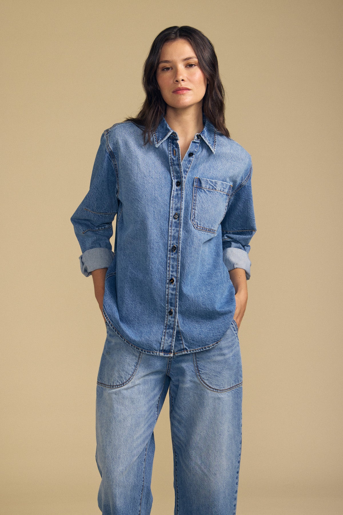 A person stands with hands in pockets, wearing the Velvet by Graham & Spencer CLEMENTINE DENIM SHIRT and loose-fitting denim jeans against a plain beige background.