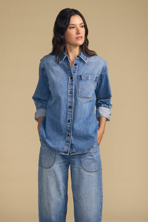 A woman stands with her hands in her pockets, wearing the CLEMENTINE DENIM SHIRT by Velvet by Graham & Spencer paired with matching wide-leg jeans against a plain beige background.