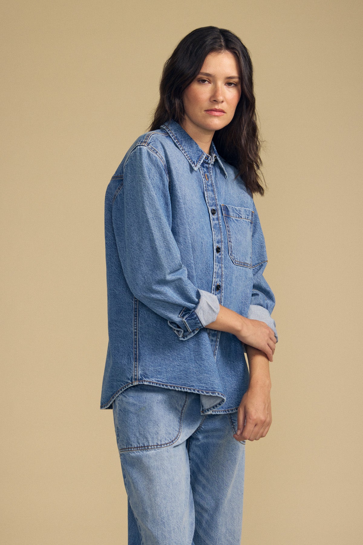 A person stands against a plain beige background wearing the CLEMENTINE DENIM SHIRT by Velvet by Graham & Spencer and matching light blue jeans, looking at the camera with a neutral expression.-41814471278785