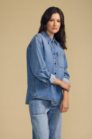 A person stands against a plain beige background wearing the CLEMENTINE DENIM SHIRT by Velvet by Graham & Spencer and matching light blue jeans, looking at the camera with a neutral expression.