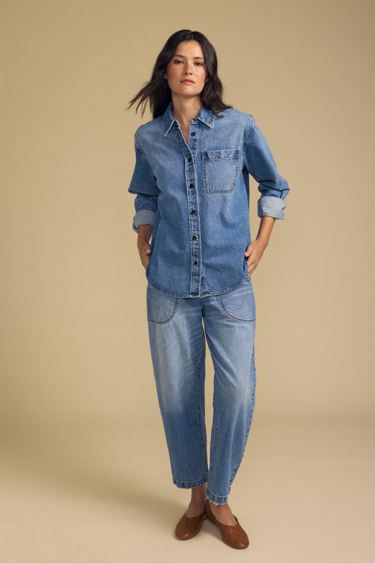 A woman stands against a beige background, wearing the CLEMENTINE DENIM SHIRT by Velvet by Graham & Spencer, paired with relaxed-fit denim jeans and brown flat shoes, her hands in her pockets.