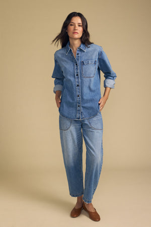 A woman stands against a beige background, wearing the CLEMENTINE DENIM SHIRT by Velvet by Graham & Spencer, paired with relaxed-fit denim jeans and brown flat shoes, her hands in her pockets.