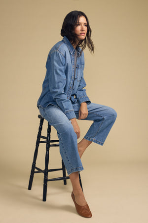 A woman wearing the CLEMENTINE DENIM SHIRT by Velvet by Graham & Spencer and jeans sits on a wooden stool against a plain beige background, looking to the side.