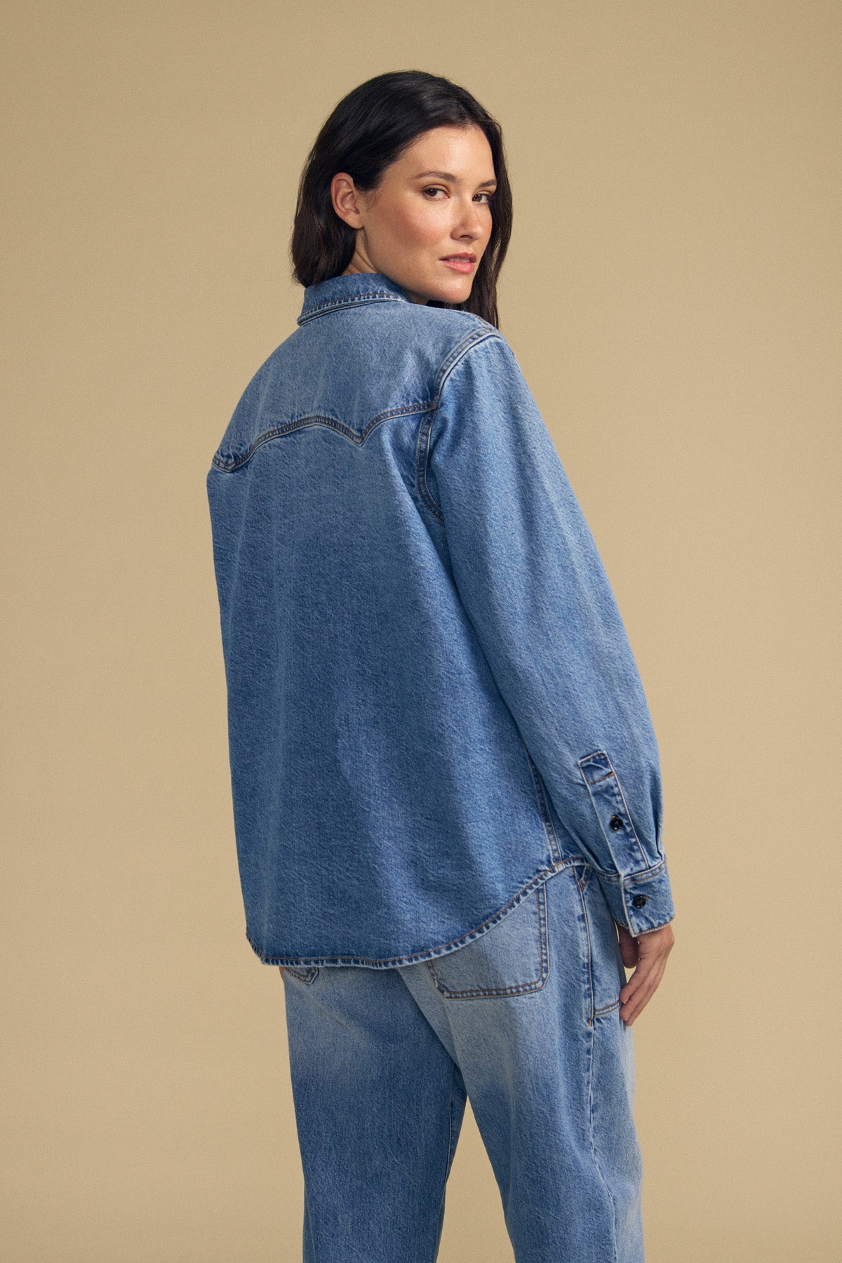 A woman stands with her back to the camera, wearing the CLEMENTINE DENIM SHIRT by Velvet by Graham & Spencer and jeans, set against a plain beige background.