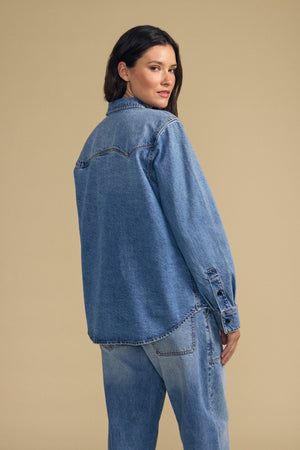 A woman stands with her back to the camera, wearing the CLEMENTINE DENIM SHIRT by Velvet by Graham & Spencer and jeans, set against a plain beige background.