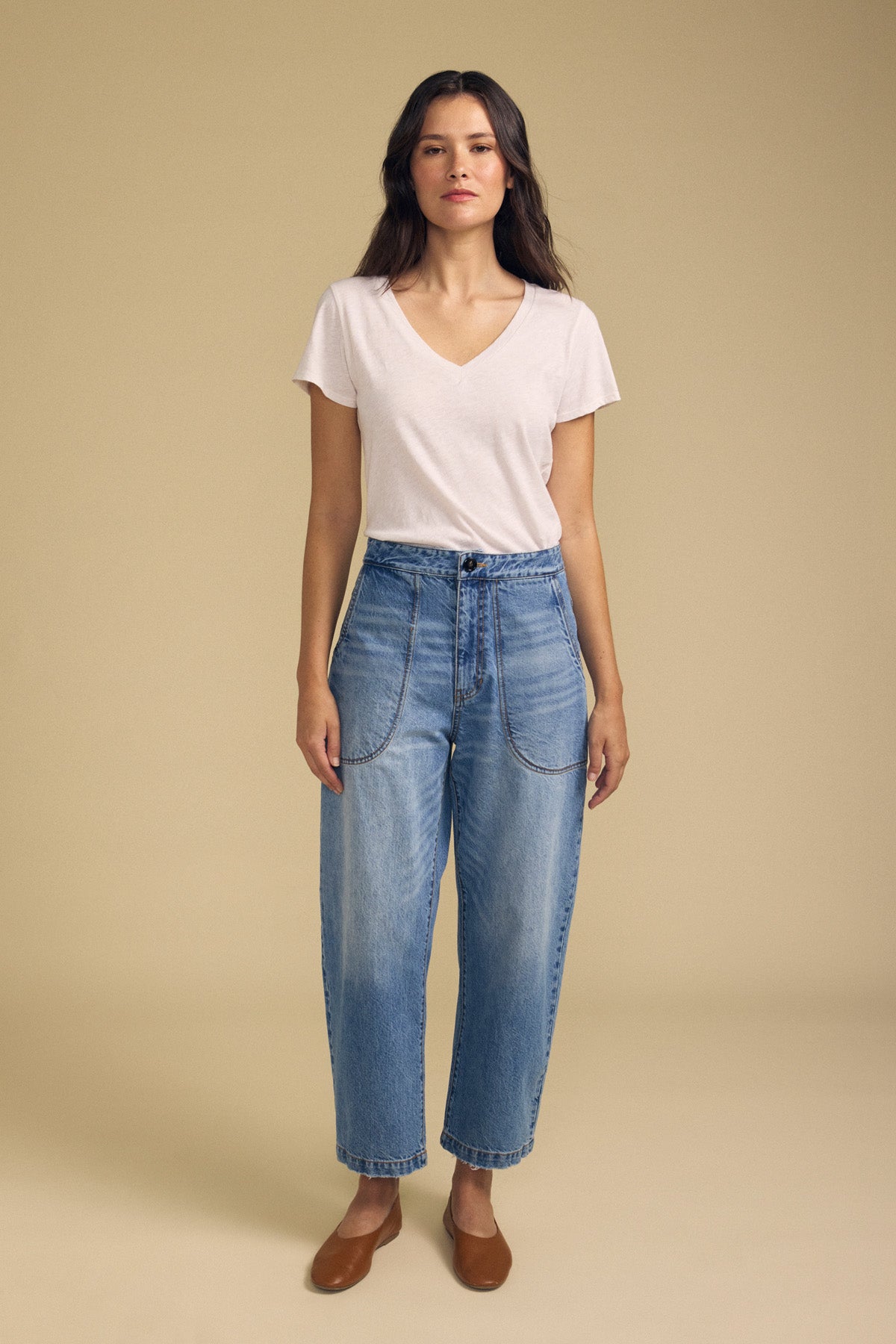 A woman stands against a plain beige background, wearing the Velvet by Graham & Spencer ESMERALDA JEAN—high-waisted blue jeans with front pockets—paired with a white V-neck T-shirt and brown flat shoes.