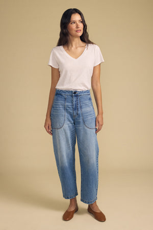 A woman wearing the ESMERALDA JEAN by Velvet by Graham & Spencer—a pair of high-waisted blue jeans with large front pockets—paired with a light pink V-neck T-shirt and brown flat shoes, stands against a beige background.