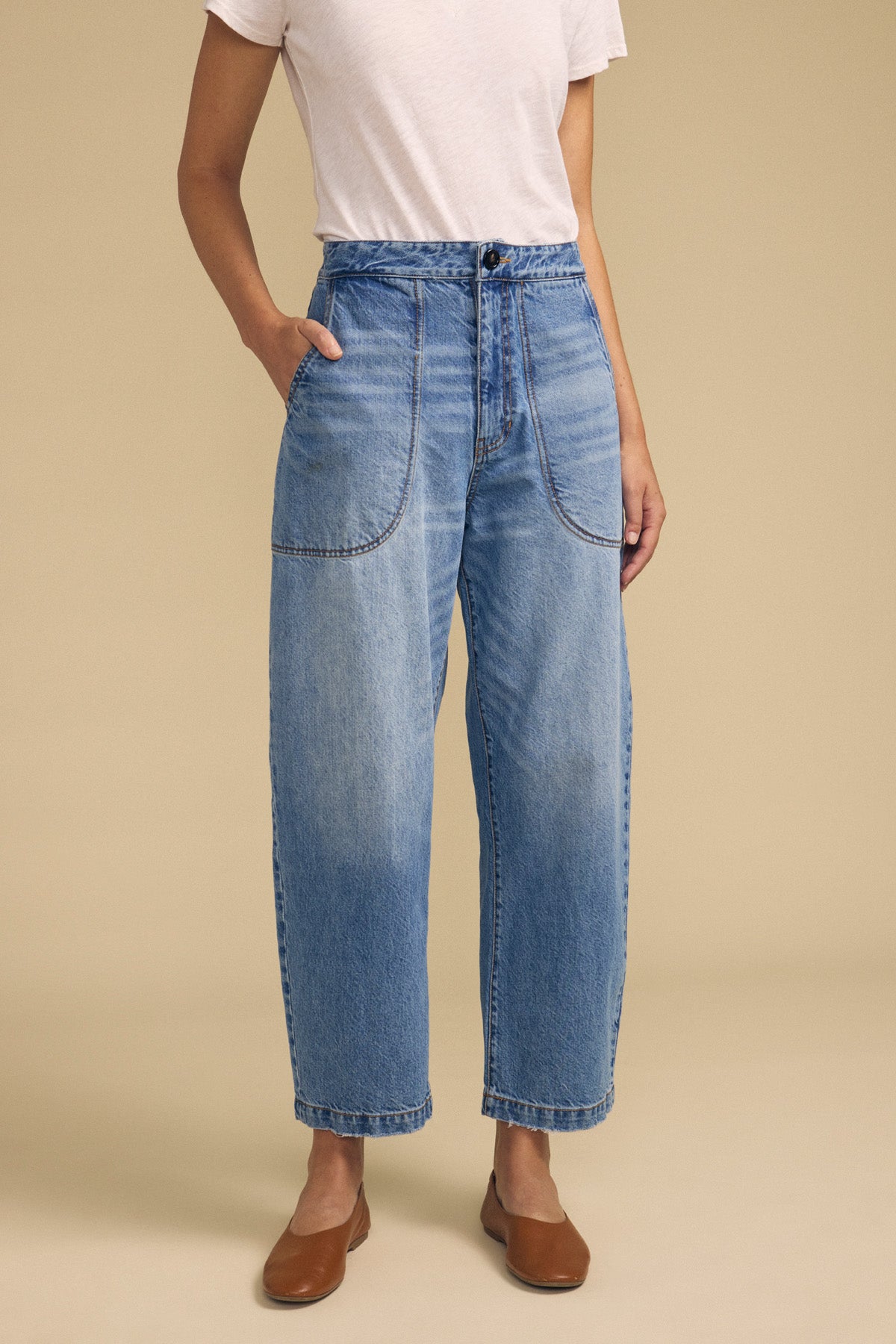 A person wearing a white t-shirt, Velvet by Graham & Spencer's ESMERALDA JEAN—a high-waisted blue wide-leg style featuring front patch pockets—and brown flats, standing against a beige background.-41815064576193