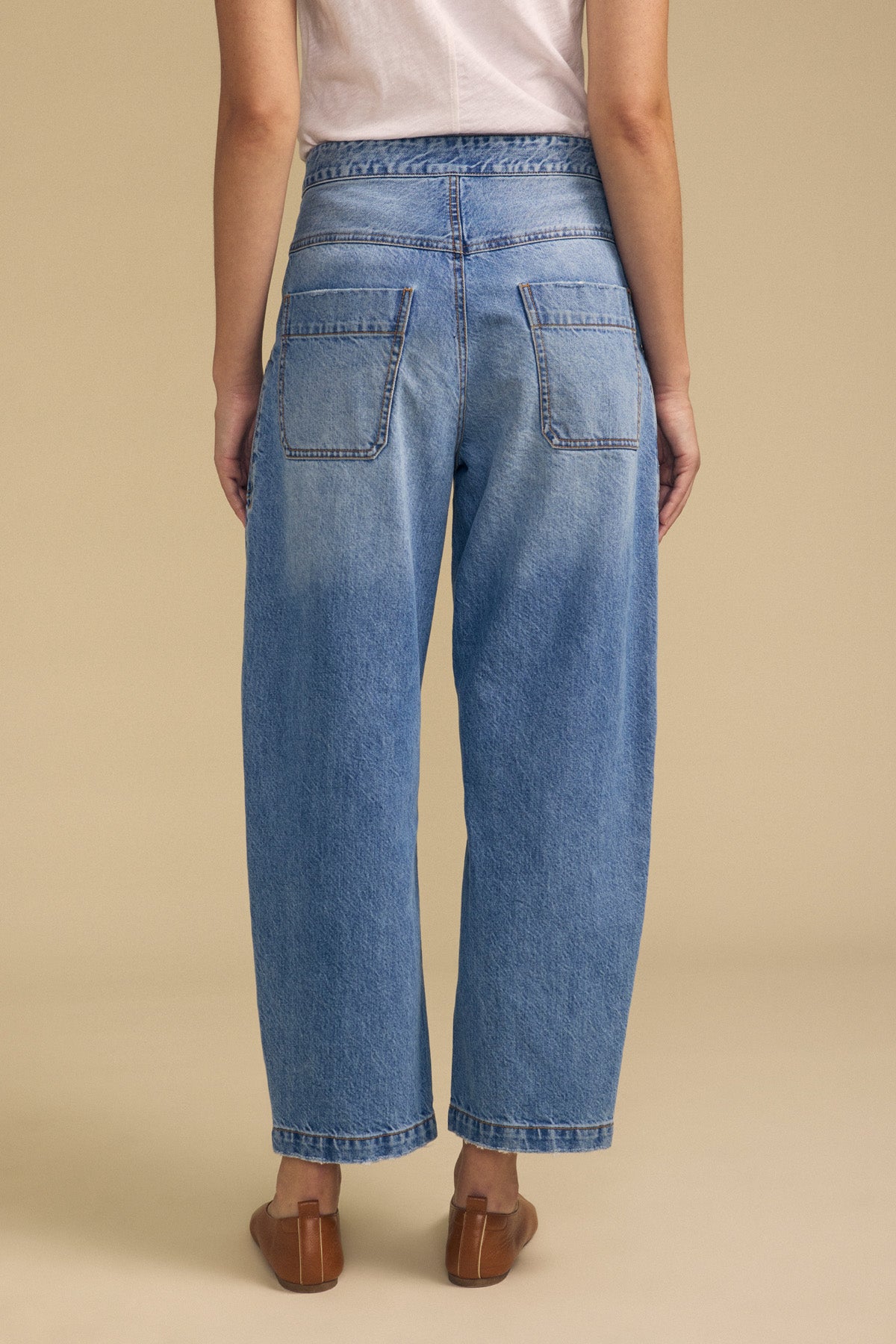 A person stands with their back to the camera, wearing a white t-shirt, Velvet by Graham & Spencer's ESMERALDA JEAN in a loose-fitting blue wash with back pockets, and brown flat shoes against a beige background.-41815064543425