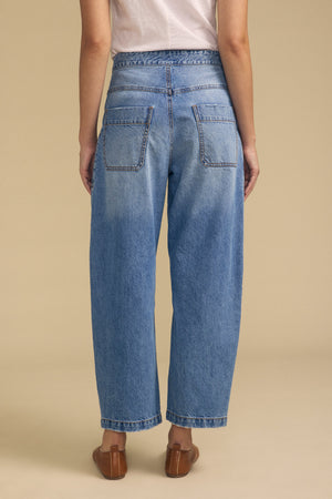 A person stands with their back to the camera, wearing a white t-shirt, Velvet by Graham & Spencer's ESMERALDA JEAN in a loose-fitting blue wash with back pockets, and brown flat shoes against a beige background.
