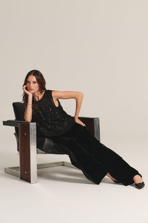 A woman reclines sideways on a modern black and chrome chair against a plain background, wearing the HILMA PANT by Velvet by Graham & Spencer and a black sleeveless sequin top.