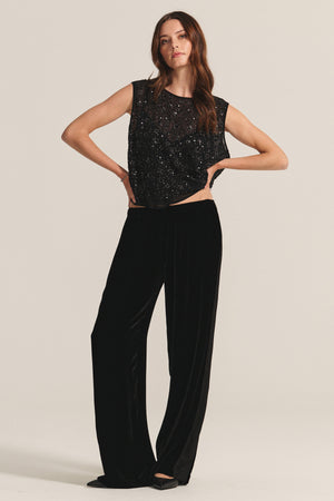 A woman stands against a plain background wearing a sleeveless black embellished top and the HILMA PANT by Velvet by Graham & Spencer, paired with black shoes.