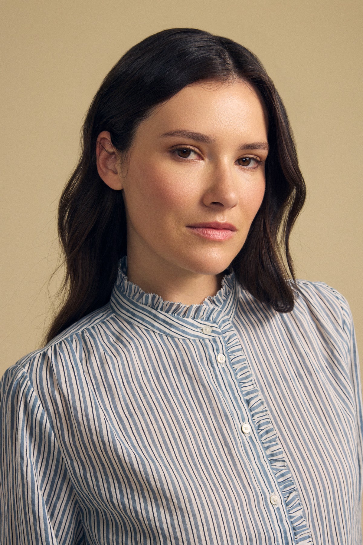 A woman with long brown hair, wearing the BETTY SHIRT by Velvet by Graham & Spencer—a blue and white striped blouse—stands in front of a plain beige background, looking calmly at the camera.