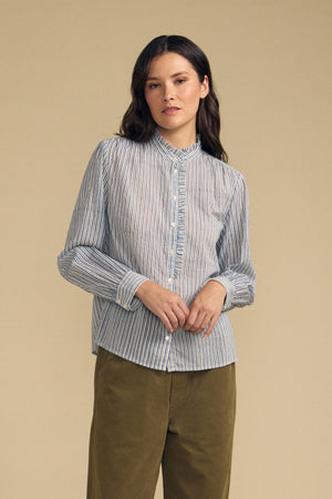 A woman wearing the BETTY SHIRT by Velvet by Graham & Spencer—a blue and white striped long-sleeve blouse—paired with olive green pants, stands against a plain beige background.
