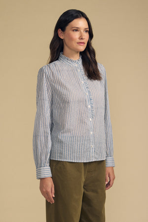 A woman stands against a beige background, wearing the BETTY SHIRT—a blue and white striped button-up with long sleeves—from Velvet by Graham & Spencer, paired with olive green pants.