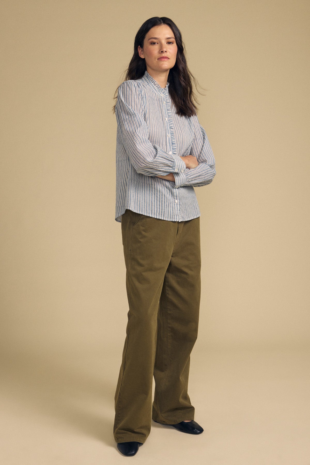 A woman with long dark hair and a neutral expression stands against a beige background, wearing the BETTY SHIRT by Velvet by Graham & Spencer—a blue striped button-up—paired with wide-leg olive green pants and black shoes.-41814381461697