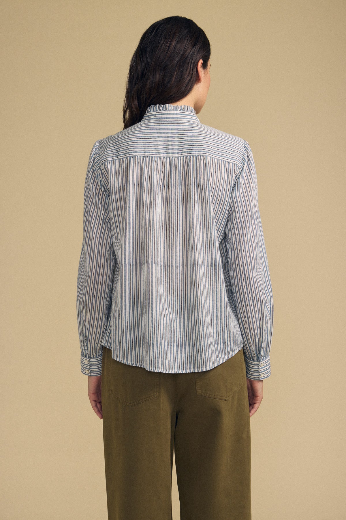 A person stands facing away, wearing the BETTY SHIRT by Velvet by Graham & Spencer—a long-sleeved, striped button-up—paired with olive green pants against a plain beige background.-41814381428929