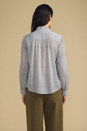 A person stands facing away, wearing the BETTY SHIRT by Velvet by Graham & Spencer—a long-sleeved, striped button-up—paired with olive green pants against a plain beige background.
