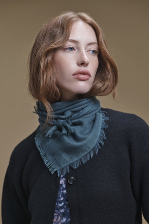 A person wears a teal triangular SUMMER CASHMERE HERRINGBONE SCARF by Velvet by Graham & Spencer, featuring a herringbone pattern and frayed edges tied around their neck.
