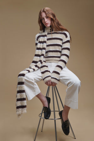 A person wearing a cozy, brown and cream striped TINA FEATHER YARN SWEATER by Velvet by Graham & Spencer with a matching scarf and white cropped trousers, sitting on a metal stool.