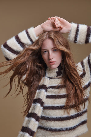 A person wearing a fuzzy, horizontally striped SALLY FEATHER YARN STRIPED SCARF and cozy accessory with their hands resting on the top of their head.