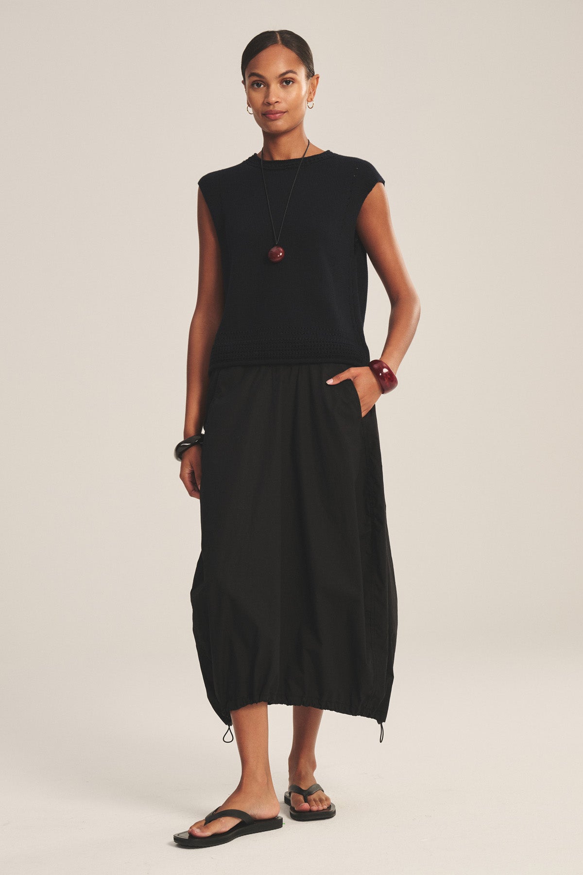 A woman stands against a plain background wearing the HOLLIS SKIRT by Velvet by Graham & Spencer, paired with a sleeveless black top, black sandals, and red jewelry. The cotton poplin skirt features an elastic waist and drawstring hem.-41038327218369
