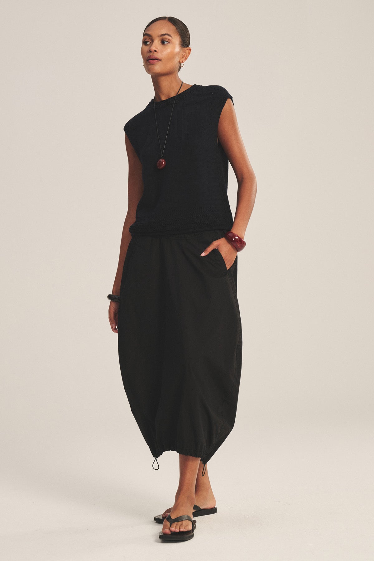 A woman wearing the HOLLIS SKIRT by Velvet by Graham & Spencer, paired with a sleeveless black top, black sandals, and red jewelry, stands against a plain light background.