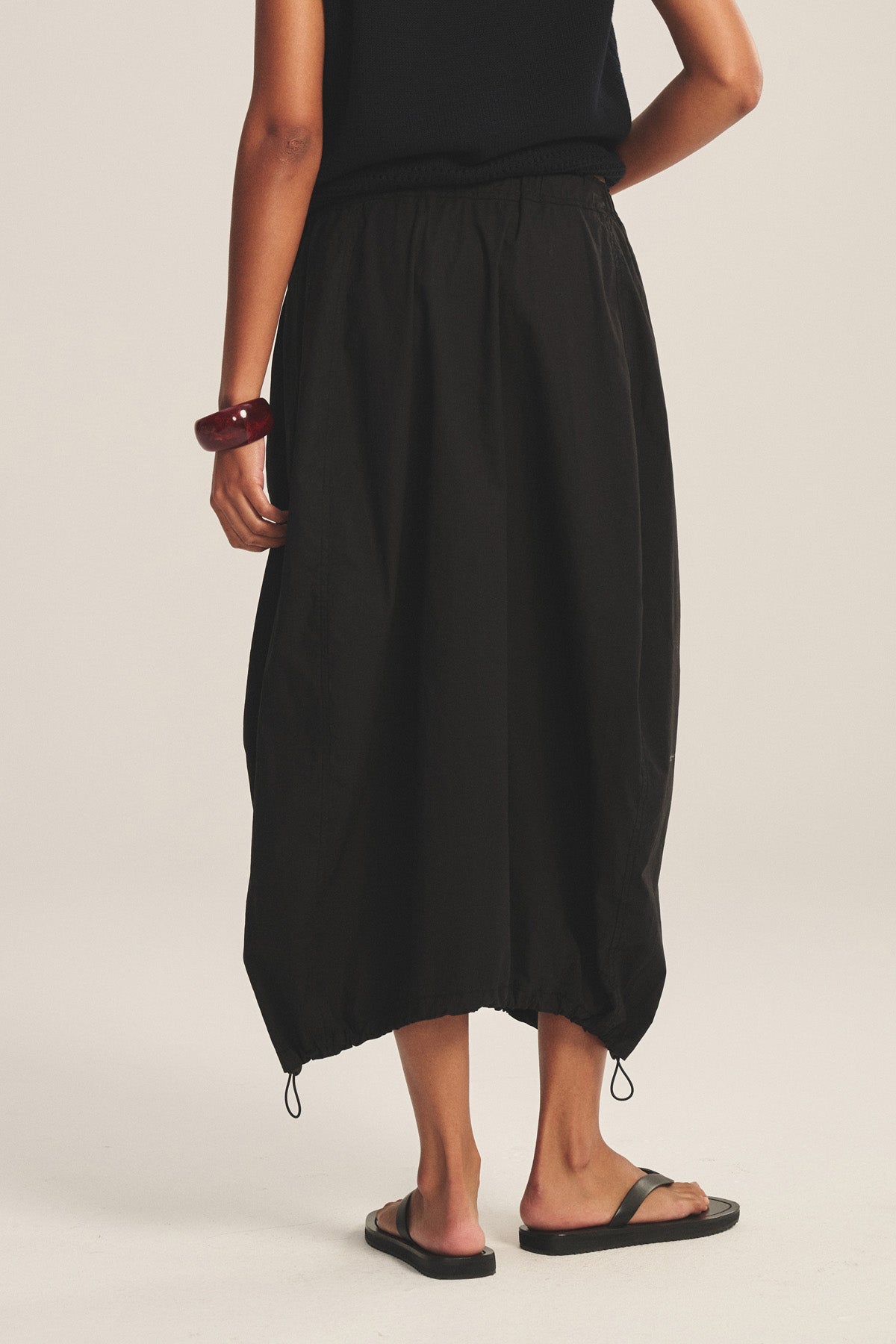 Person wears a black sleeveless top, black sandals, chunky brown bracelet, and the HOLLIS SKIRT by Velvet by Graham & Spencer—a loose cotton poplin skirt with elastic waist and drawstring hem—standing against a plain background.