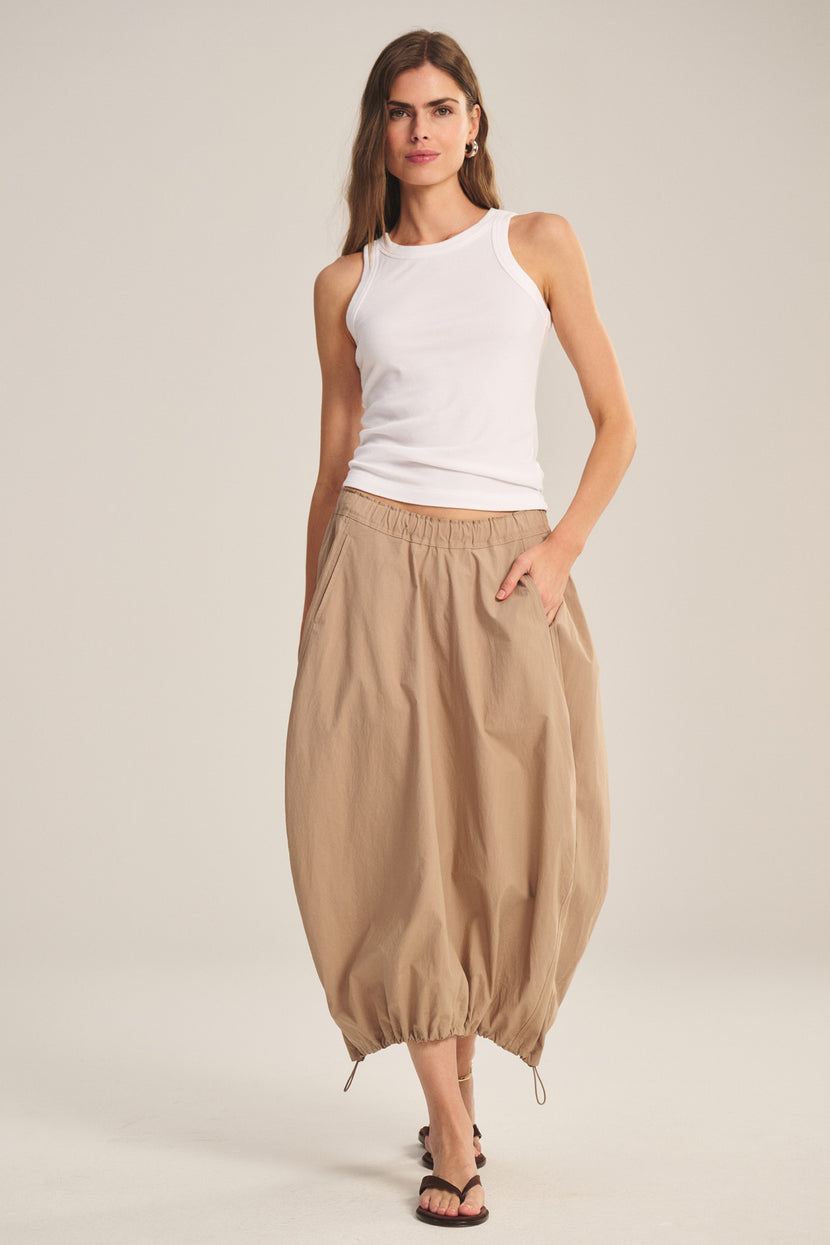 A woman stands against a plain background wearing a white sleeveless top, black sandals, and the HOLLIS SKIRT by Velvet by Graham & Spencer—a beige cotton poplin skirt with an elastic waist.