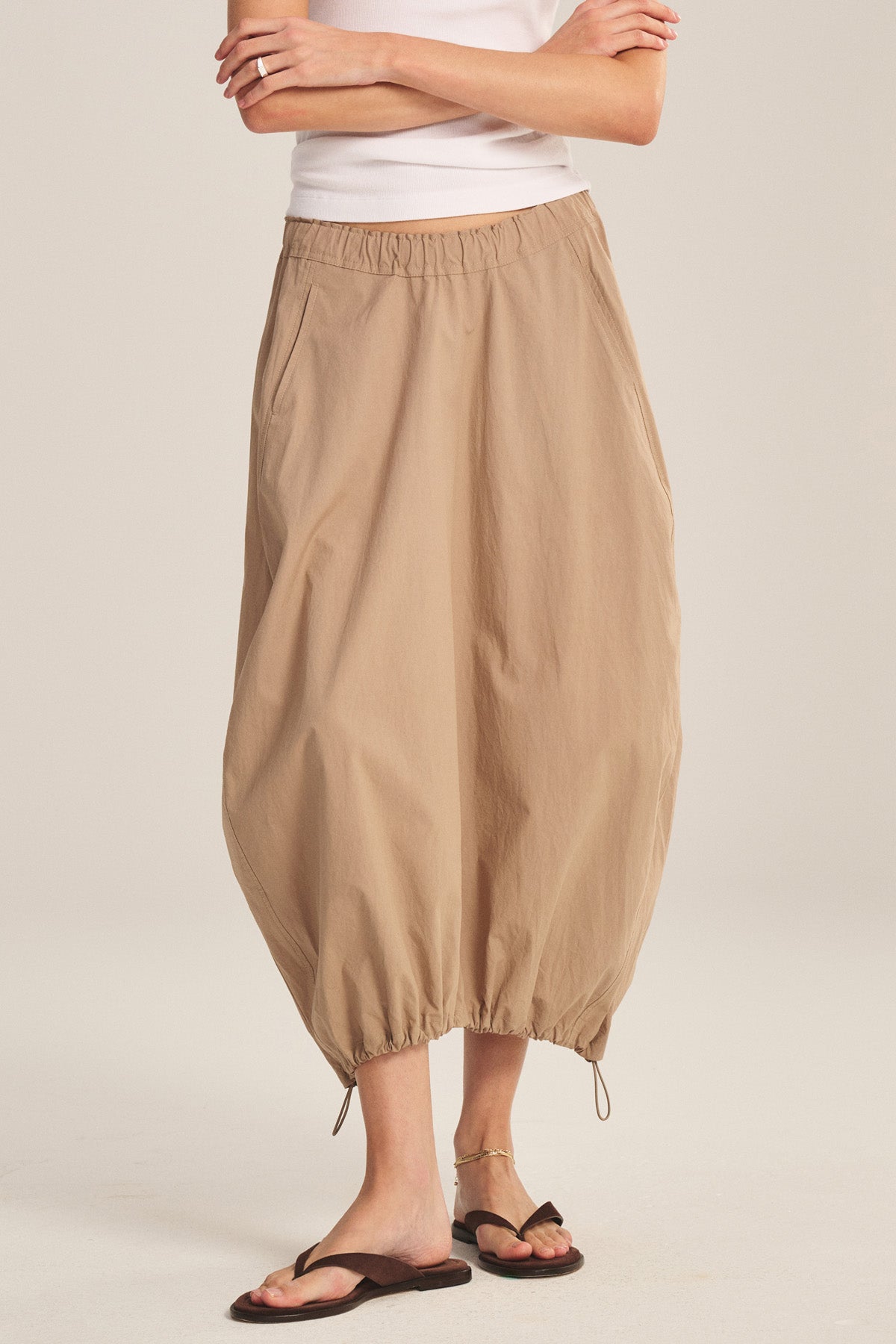 A person wearing the Velvet by Graham & Spencer HOLLIS SKIRT in tan with an elastic waist and cuffs, paired with a white top and black sandals, standing with arms crossed.-41338481246401